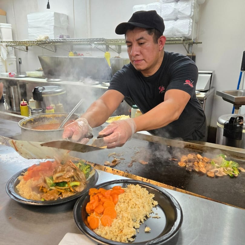 A chef at Sake Express cooking Japanese hibachi-style food on a griddle, preparing two plates.