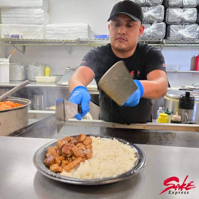 A Sake Express chef in blue gloves prepares a plate of chicken and rice in a bustling kitchen.