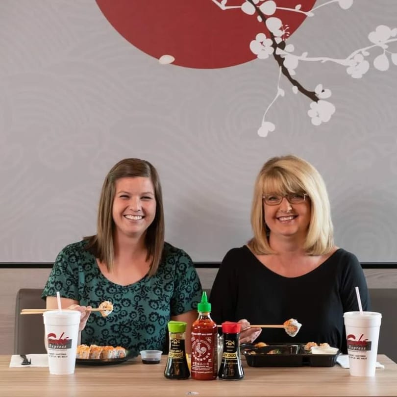 Two smiling women enjoying sushi and drinks at a table in Sake Express.