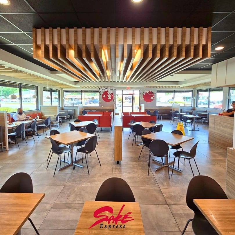 Spacious Sake Express dining room with wooden tables, black chairs, red booths, and decorative ceiling panels.