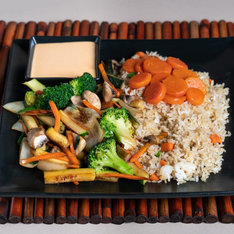Japanese hibachi meal with fried rice, mixed vegetables, and a side of sauce on a black plate.