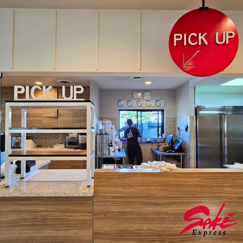 A Sake Express restaurant interior with a pick-up counter, a worker, and a red circular pick-up sign.