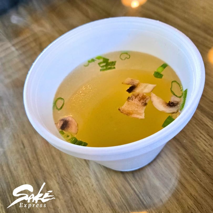 A white bowl of clear Japanese soup with sliced mushrooms and green onions on a wooden table.