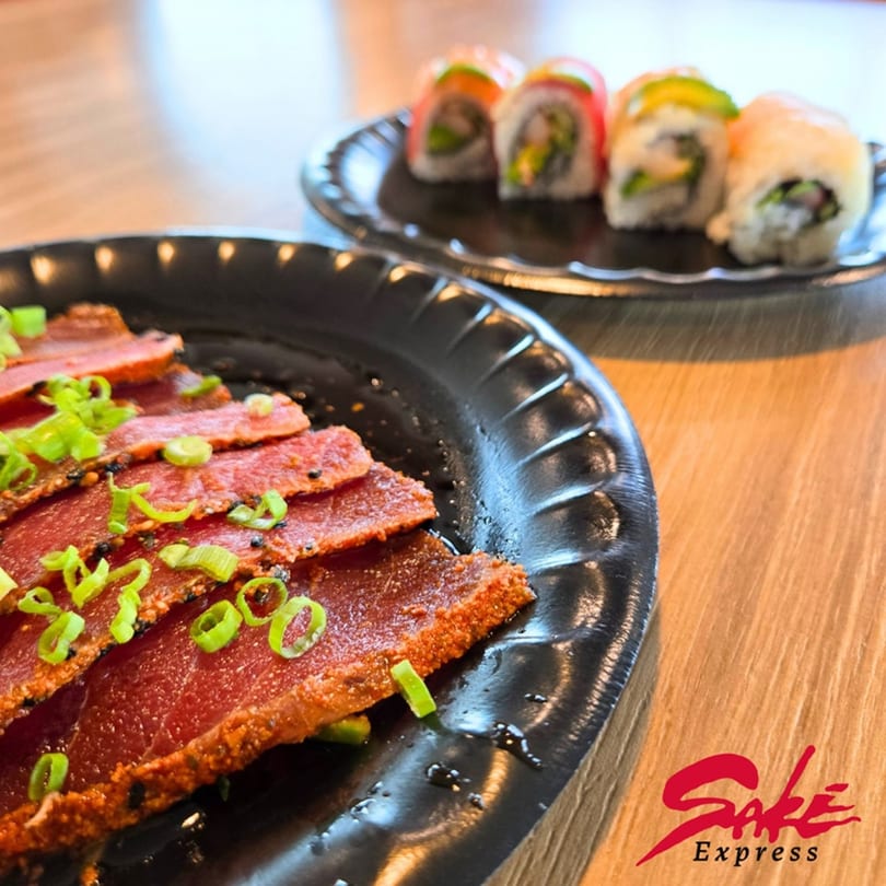 Seared tuna sashimi with green onions and sesame seeds, alongside sushi rolls on black plates.