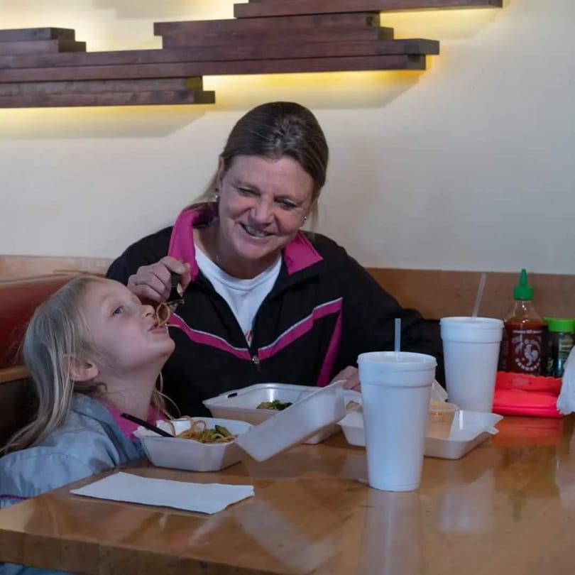 A smiling woman feeds noodles to a young girl at a wooden table in Sake Express.