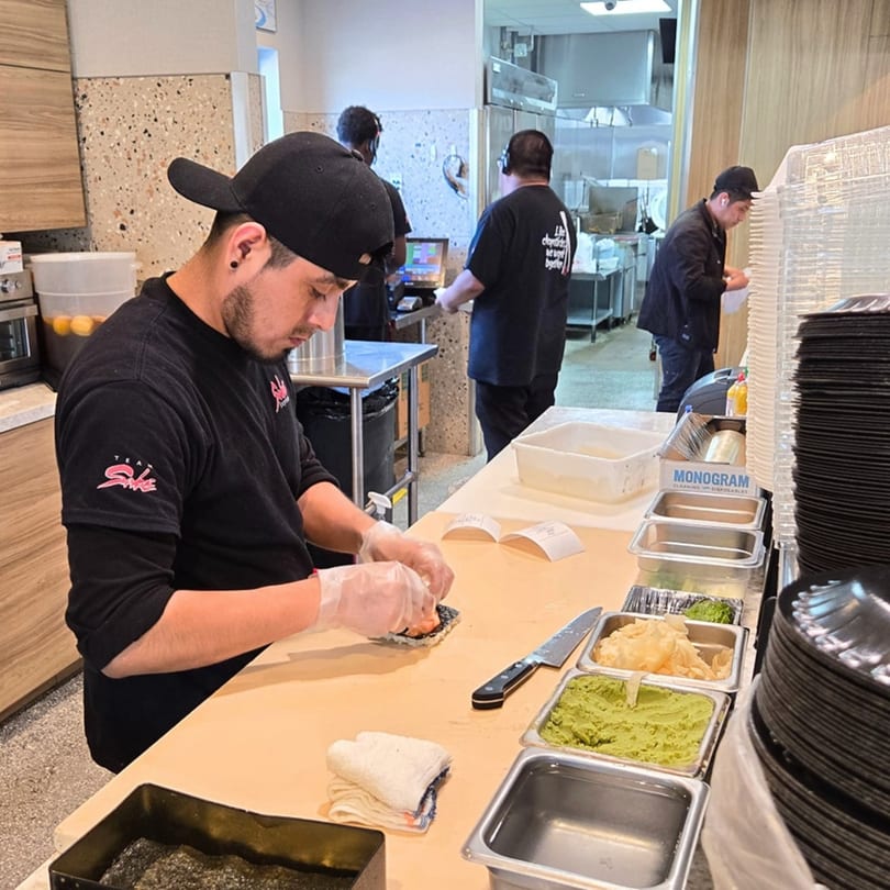A Sake Express chef in a black shirt and cap prepares sushi behind a counter with ingredients.
