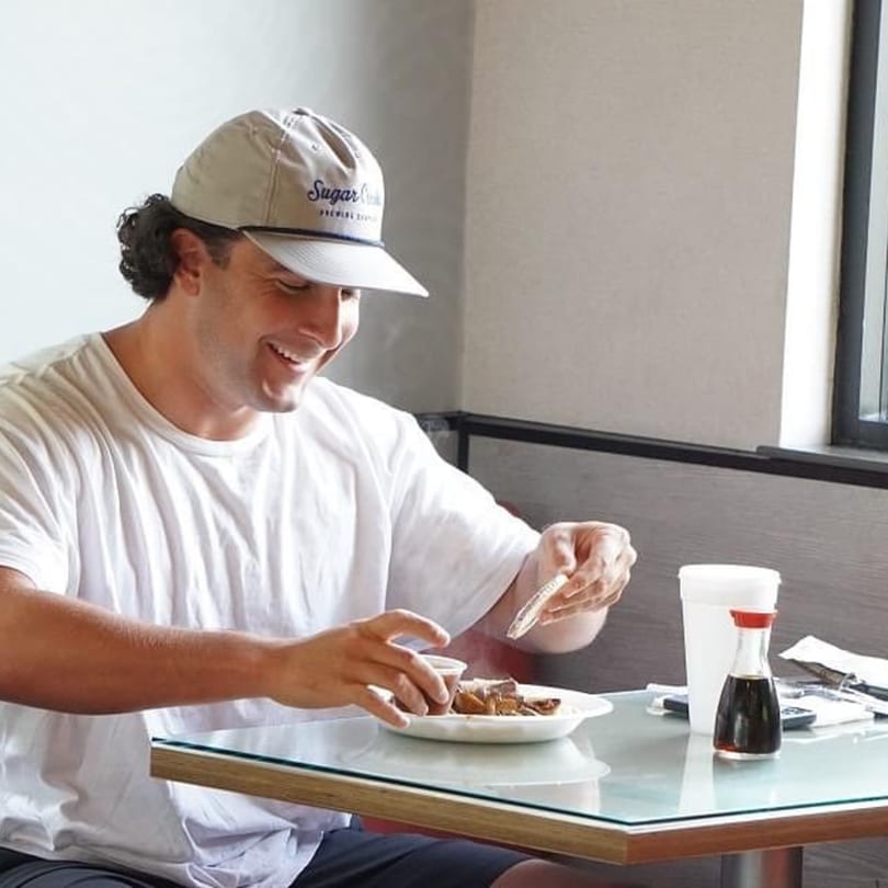 A man in a white t-shirt and cap smiles while eating Japanese food at Sake Express.
